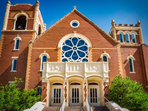 Historic brick church with a large rose window, bell tower, and decorative entrance surrounded by greenery.