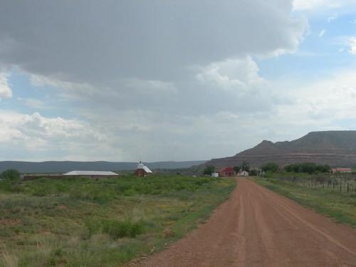 A dirt road leads through green fields towards a distant farm and mountains under a cloudy sky.