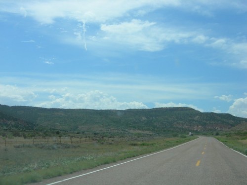 A scenic view of a winding road leading through green hills under a blue sky with scattered clouds.