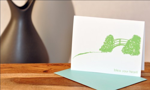 A greeting card with a green illustration of a bridge and trees, resting on a wooden surface next to a vase.
