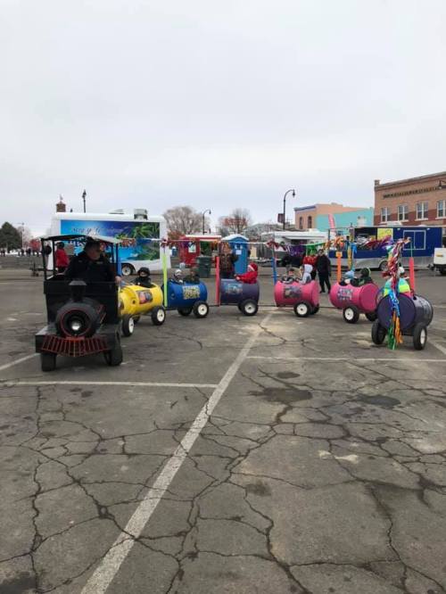 A colorful train ride for children parked in a lot, with a small engine and several vibrant carriages.
