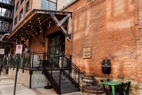 A brick building with string lights, a staircase, and outdoor seating, featuring wooden barrels and a sign on the wall.
