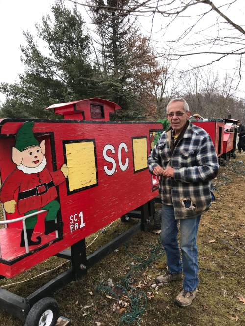 An older man in a plaid jacket stands beside a colorful, painted train with a festive elf design.
