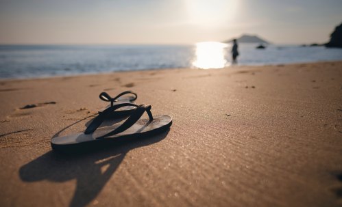 A black flip-flop rests on sandy beach, with a person walking by the water and a sunset in the background.