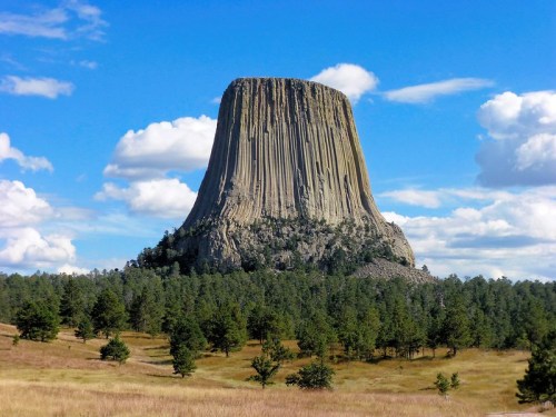 A towering rock formation rises above a grassy landscape, surrounded by trees under a blue sky with fluffy clouds.