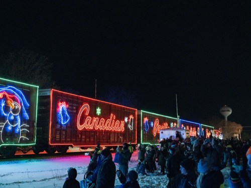 A brightly lit train with festive decorations passes by a crowd of people in a snowy night setting.