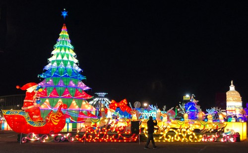 Colorful holiday lights illuminate a large Christmas tree and festive decorations at night, with a person walking nearby.