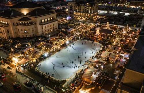 Aerial view of a festive ice skating rink surrounded by holiday lights and market stalls at night.
