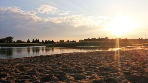 Sunset over a calm river, with sandy shore and soft clouds in the sky, creating a serene and peaceful atmosphere.