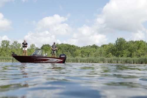 Two people stand on a boat in a calm lake, surrounded by lush greenery and a partly cloudy sky.