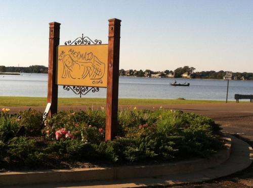 Sign for "The Mermaid Cafe" near a lake, surrounded by flowers and greenery, with a boat in the background.
