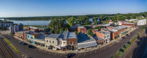 A panoramic view of a small riverside town with historic buildings, trees, and a calm river in the background.