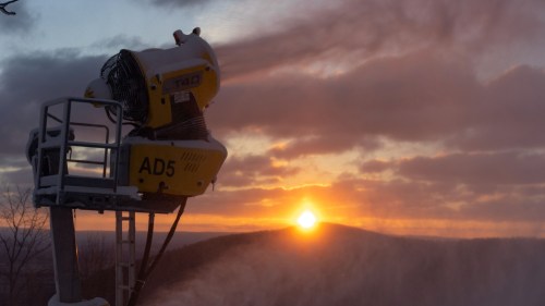 A snowmaking machine stands against a sunset backdrop, with clouds and a snowy landscape in the foreground.