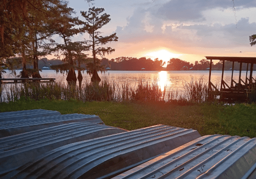 Sunset over a calm lake, with metal boats lined up on the grass and trees reflecting in the water.