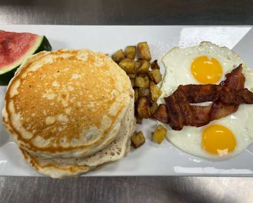 A plate of pancakes, crispy bacon, fried eggs, and diced potatoes, with a slice of watermelon on the side.