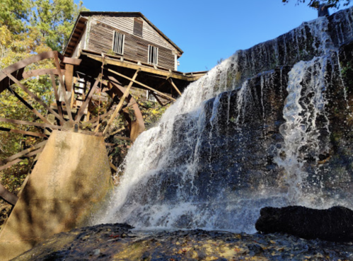 A historic watermill with a large wooden wheel, cascading water, surrounded by trees and a clear blue sky.