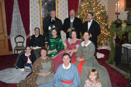 A group of people in 19th-century attire poses in a festive room decorated for the holidays, with a Christmas tree.