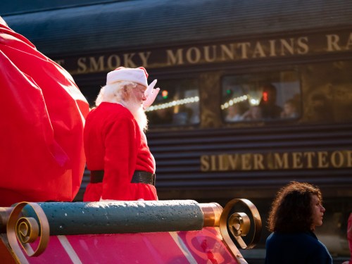 Santa Claus stands on a festive float, with a train in the background, during a holiday parade.