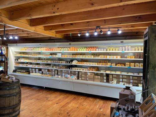 A well-organized cheese shop display with various cheeses and products on shelves, wooden beams, and a rustic floor.