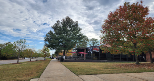 A sidewalk lined with trees, showcasing fall colors, leads to a restaurant with outdoor seating under a cloudy sky.
