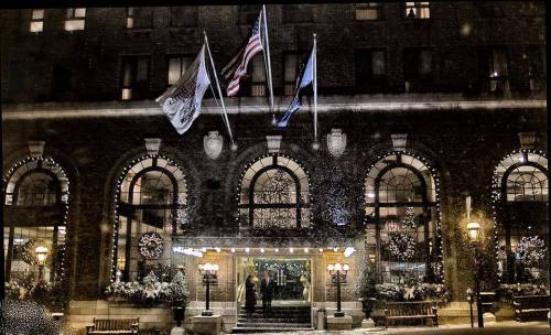 A historic hotel entrance adorned with holiday decorations, flags, and snow gently falling in the evening light.