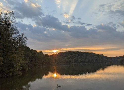 Sunset over a calm lake, with rays of light breaking through clouds and reflecting on the water's surface.