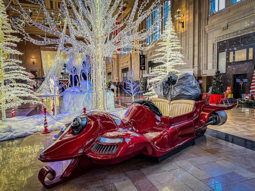 A festive red sleigh with a large bag sits in a beautifully decorated hall with sparkling trees and holiday lights.