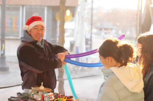 A man in a Santa hat twists balloons while two people watch, enjoying a festive atmosphere.