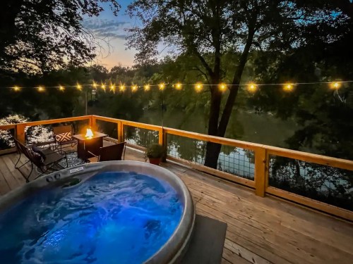 A serene deck with a hot tub, string lights, and a view of a river at sunset, surrounded by trees.