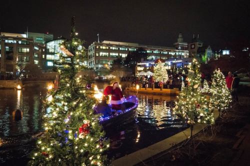 A festive scene with a Santa figure on a boat, surrounded by decorated trees and a crowd by the water at night.