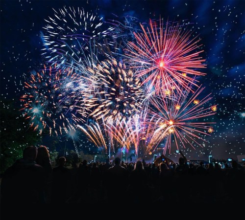 A vibrant fireworks display lights up the night sky, with silhouettes of people watching in the foreground.