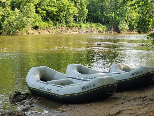 Three inflatable rafts are tied to the shore of a calm river, surrounded by lush green trees.