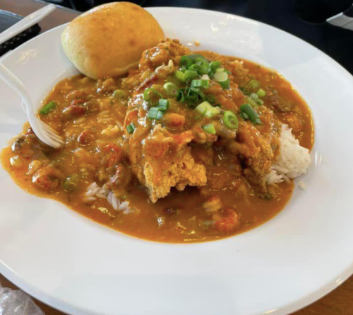 A plate of fried chicken topped with gravy, served over rice and garnished with green onions, alongside a bread roll.