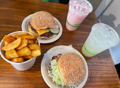 Two sesame seed burgers with toppings, a side of potato wedges, and two drinks on a wooden table.