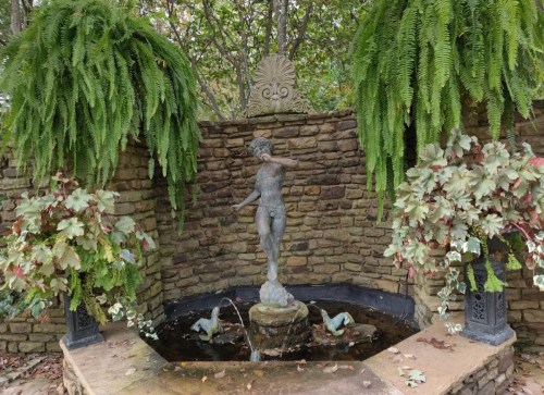 A stone fountain featuring a statue of a boy surrounded by lush ferns and colorful plants in a serene garden setting.