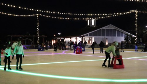 A lively ice skating rink at night, illuminated by colorful lights, with skaters and a building in the background.