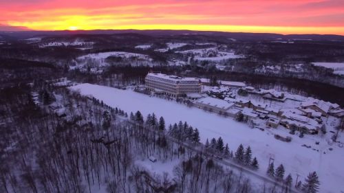Aerial view of a snowy landscape at sunset, featuring a large building surrounded by trees and hills.