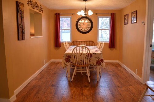 Cozy dining room with a wooden table, chairs, and a large wall clock, featuring warm yellow walls and natural light.