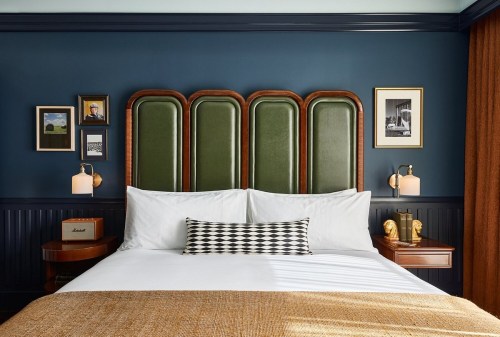 A stylish hotel bedroom featuring a green upholstered headboard, white bedding, and framed artwork on dark blue walls.