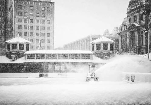 A snow-covered cityscape with a snowplow clearing a path amidst heavy snowfall and tall buildings in the background.