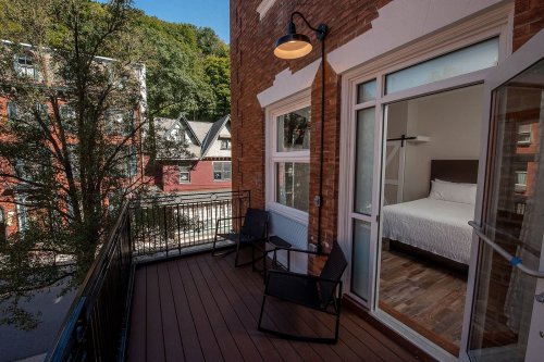 A balcony with two chairs overlooks a street, featuring a view of nearby buildings and a room with a bed inside.