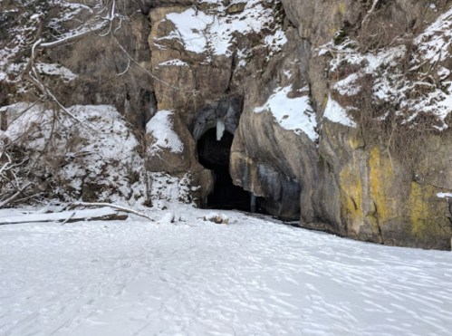 A snowy landscape featuring a large rock cave entrance surrounded by rocky cliffs and sparse vegetation.