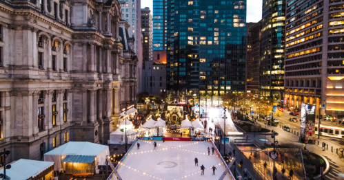 Aerial view of a city ice skating rink surrounded by buildings, with festive lights and decorations at dusk.