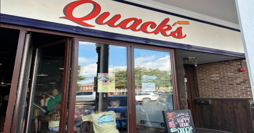 Exterior of Quack's café with large windows, people inside, and a sign indicating it's open. Bright and inviting atmosphere.