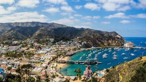 A scenic view of a coastal town with boats in the harbor, surrounded by mountains and blue skies.