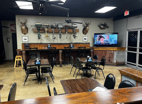 Interior of a rustic restaurant with wooden tables, black chairs, and a TV displaying a cartoon, adorned with mounted deer heads.
