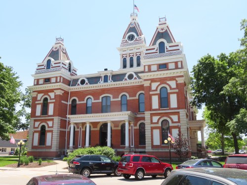 Historic brick courthouse with ornate architecture, clock tower, and surrounding trees and parked cars on a sunny day.