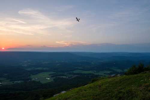 A small plane flies over a scenic valley at sunset, with rolling hills and fields below.