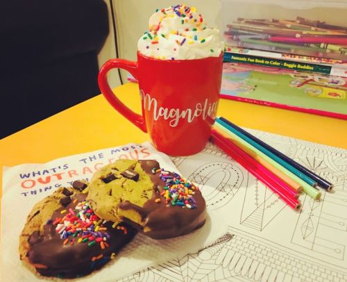 A red mug with whipped cream and sprinkles, cookies on a napkin, and colorful pens beside a coloring book.