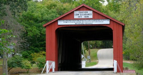 A red covered bridge with a low clearance sign, surrounded by green trees and a winding road.